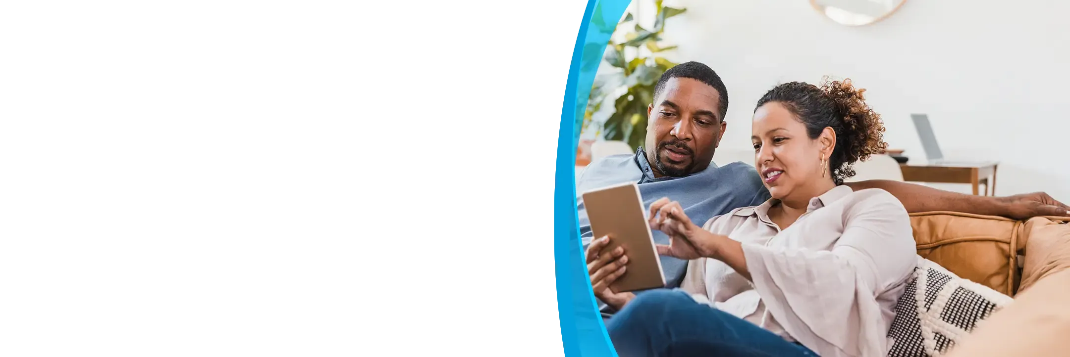 A couple relaxes on a couch together browsing a tablet in a bright living room.